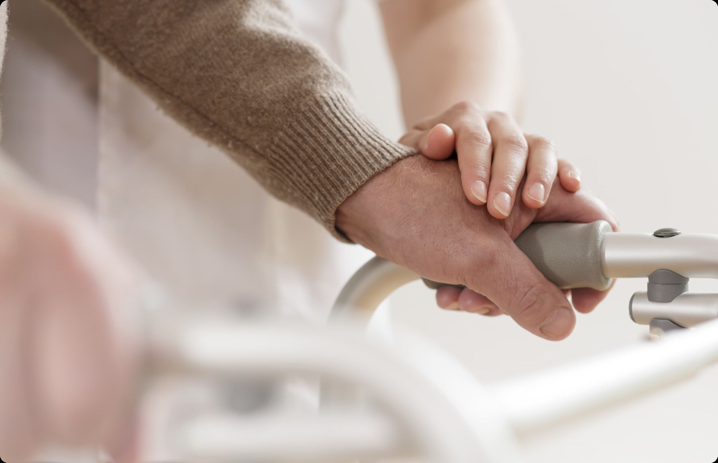 Die Hand eines älteren Menschen hält den Griff eines Rollators mit Unterstützung einer zweiten, fremden Hand.